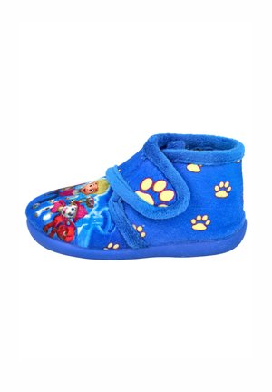 Zapatilla de casa de forro polar azul con una correa de velcro, con estampados coloridos de personajes y huellas amarillas. Material suave y texturizado con punta redondeada.