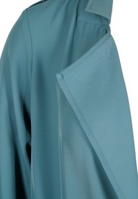 Tessuto blu-verde con un primo piano di una manica che presenta un dettaglio a flap ripiegato e cuciture visibili su un materiale liscio.
