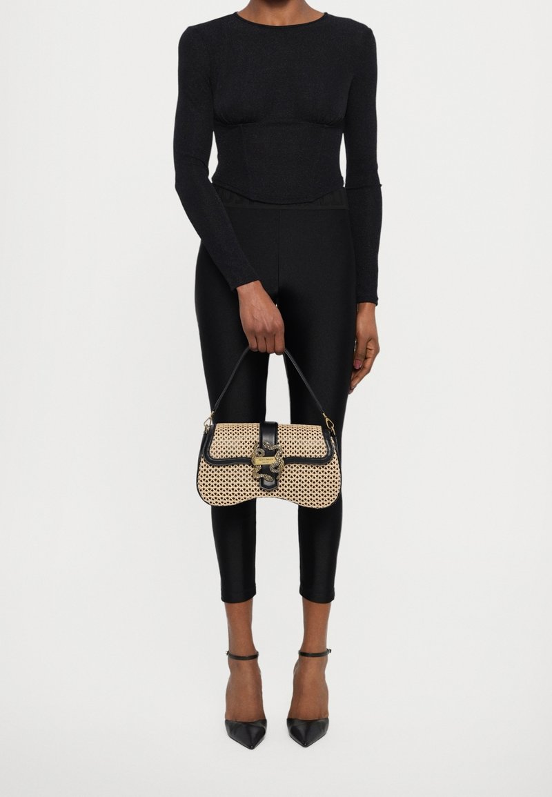Personne portant un haut noir ajusté à manches longues et un pantalon, tenant un sac tissé beige avec garniture noire et fermoir, portant des talons pointus noirs.