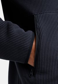 Giacca a coste scura con tasca con zip e linguetta in panno. La texture è morbida e il tessuto ha un motivo uniforme.