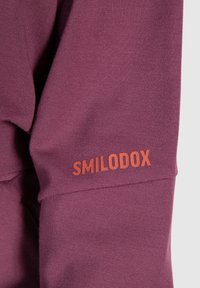 Gros plan d'une manche en tissu grenat avec un logo orange "SMILODOX" imprimé près de la couture.