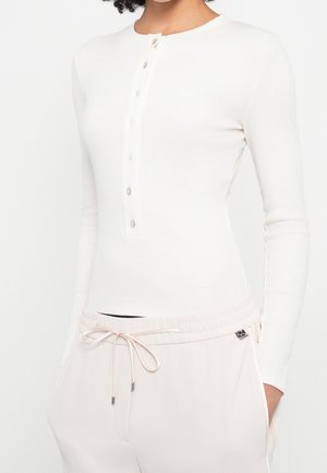 Personne portant un t-shirt henley blanc ajusté à manches longues avec des boutons et un pantalon rose clair à cordon avec les mains dans les poches.