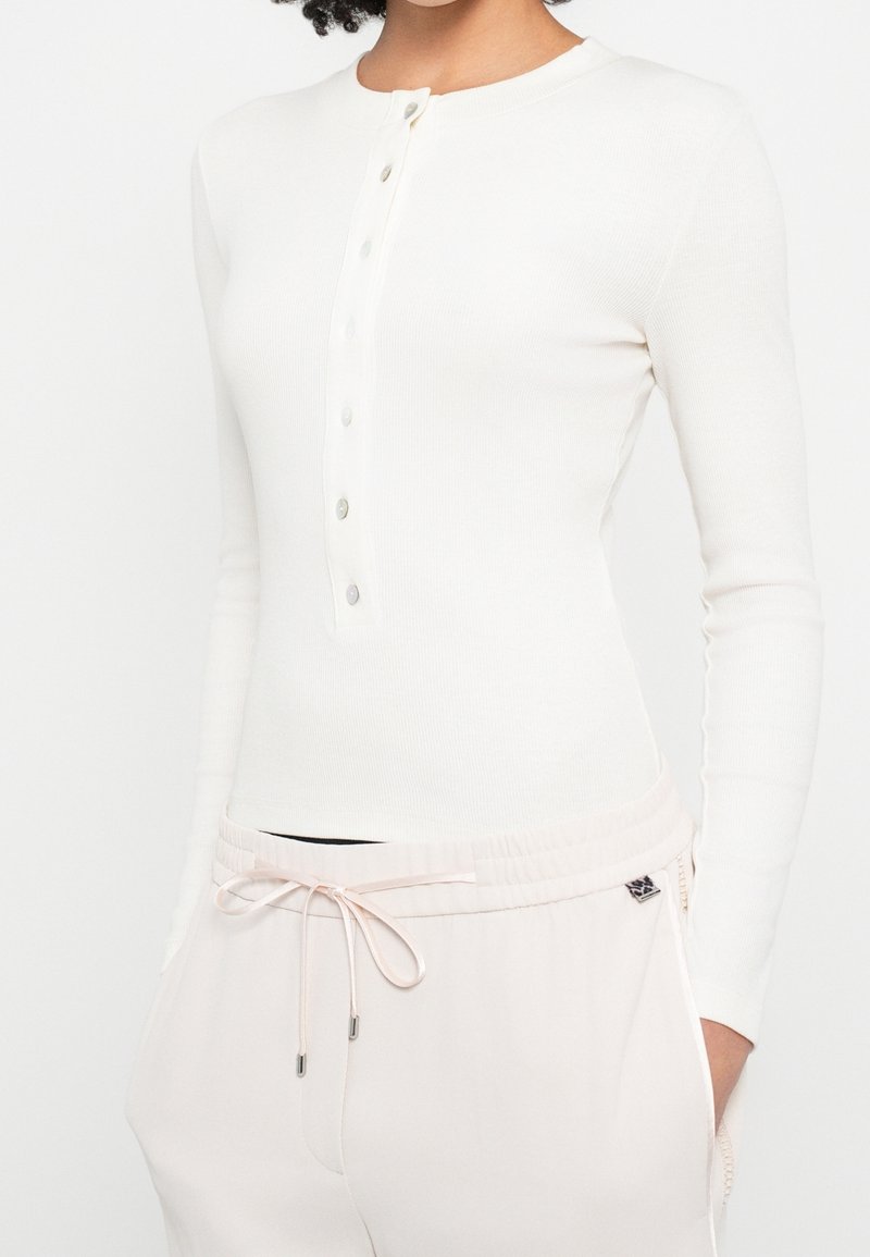 Personne portant un t-shirt henley blanc ajusté à manches longues avec des boutons et un pantalon rose clair à cordon avec les mains dans les poches.