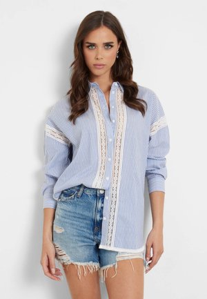 Chemise bleue à rayures avec une finition en dentelle blanche, devant à boutons et manches retroussées, associée à un short en denim déchiré.