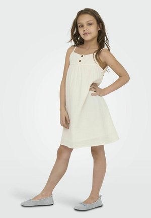 Robe A-line crème clair avec de fines bretelles, ornée de trois boutons marron sur le buste. Associée à des ballerines grisâtres. Texture lisse.