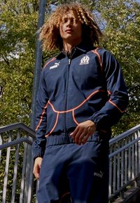 Mornarski športni trenirka z oranžnimi poudarki, vsebuje jakno na zadrgo in hlače z elastiko v pasu, izdelana iz lahkotnega materiala, označena z logotipom Puma.