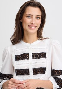 Blouse blanche avec des accents en dentelle noire et fermeture à boutons. Présente des manches bouffantes et une bordure à motifs avec des découpes délicates tout au long.