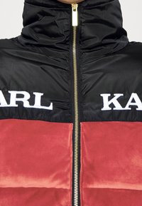 Prošívaná bunda s černou horní částí a červenou spodní částí, se zlatým zipem a vyšívanými bílými logy "KARL" a "KA".