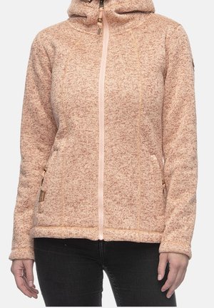 Chaqueta de forro polar con cremallera y capucha en rosa claro con textura, llevada por una persona con pantalones negros, manos apoyadas a los lados sobre un fondo liso.