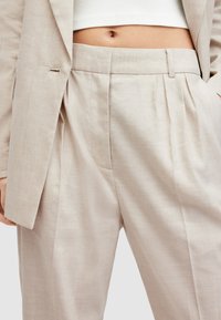 Beżowe lniane spodnie z plisami, z dopasowanym paskiem i kieszenią. Gładka faktura, subtelnie pomarszczone wykończenie, część garnituru.