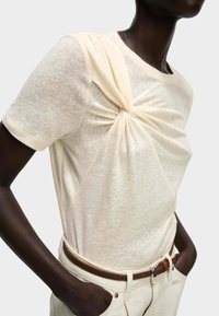 T-shirt crema a maniche corte con dettaglio frontale torsionato, texture morbida e vestibilità rilassata, abbinato a pantaloni bianchi e una cintura marrone.