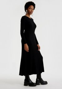 Schwarzes, geripptes Langarmkleid mit tailliertem Oberteil und ausgestelltem Saum, getragen mit massiven schwarzen Plateaustiefeln und großen herzförmigen Ohrringen.