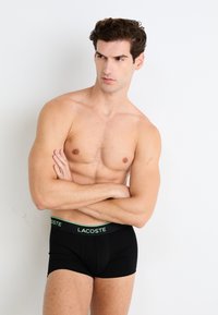 Zwarte boxershorts met een groene "LACOSTE" tailleband gedragen door een man met gekruiste armen. Glad materiaal, aansluitend ontwerp, minimale detailing.