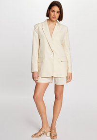 Blazer entallado color crema con sutiles lunares, cierre de un solo botón y dos bolsillos frontales, combinado con pantalones cortos a juego y tacones de tiras.