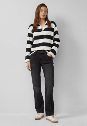 Donna con capelli castani che indossa una maglia a maniche lunghe a righe bianche e nere, jeans neri, sneakers bianche, in piedi davanti a uno sfondo grigio uniforme.