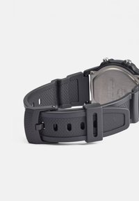 Pulseira de relógio em borracha preta com acabamento texturizado, apresentando uma fivela de metal e múltiplos buracos de ajuste, mostrando o verso da caixa do relógio.
