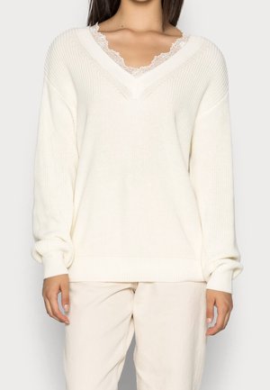 Kvinde iført en løs hvid V-hals sweater lagdelt over en bluse med blonderkant og lyse bukser, vist fra skuldre til hofter.