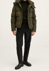 Olijfgroene donsjas met grote doorgestikte delen, ritssluiting en drukknopen, twee zakken aan de voorkant, gecombineerd met zwarte cargobroek en zwarte laarzen.