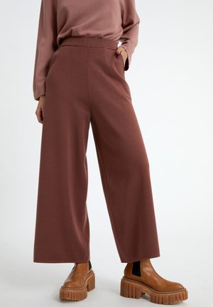 Personne portant un pantalon large marron, un haut à manches longues assorti et des bottes plateformes marron épaisses, debout avec une main dans la poche.