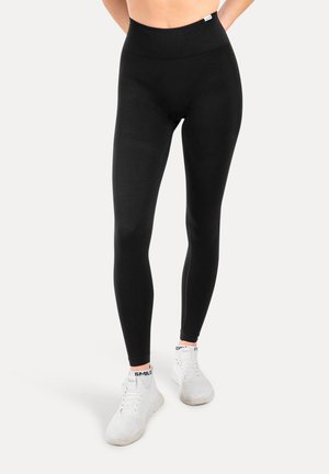 Hoch taillierte schwarze Leggings aus dehnbarem Material. Sie verfügen über eine glatte Textur und ein enganliegendes Design, kombiniert mit weißen Sportschuhen.