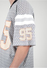 Persona con camisa de color gris claro con estampado, con un gran número 95 perforado con contorno naranja en la manga y dos franjas horizontales blancas encima.