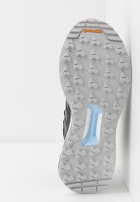 Suela de zapatilla de trail running en gris y azul con un patrón de relieve agresivo y la marca Continental, inclinada contra una pared y suelo blancos.