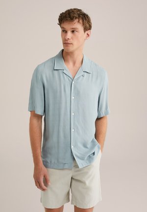 Camisa de manga corta de color azul claro con cuello y cierre de botones en la parte delantera. Fabricada con un tejido suave y texturizado, de corte relajado. Combinada con pantalones cortos claros.