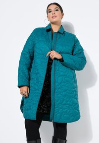 Teal quiltet jakke med lynlås, der har krave og struktureret mønster. Bæres over en sort pailletkjole og sorte leggings.