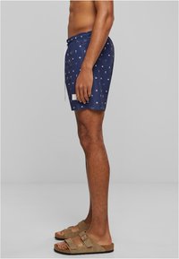 Shorts de bain navy avec un motif nautique répété, taille élastique avec cordon de serrage, et une étiquette blanche sur l'ourlet. Portés avec des sandales brunes.