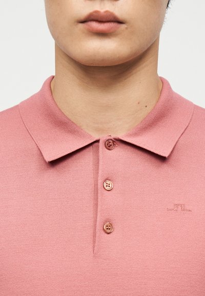 Κοντινή λήψη ενός ατόμου που φοράει ένα ροζ πόλο πουκάμισο με τρία κουμπιά και ένα μικρό κεντημένο λογότυπο στο στήθος.