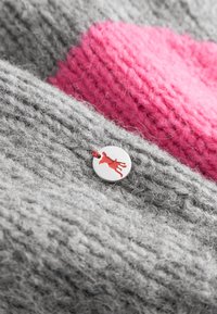 Close-up van gebreide stof met grijs en roze garen en een ronde metalen label met de afbeelding van een rode hertensilhouet, bevestigd met rood garen.