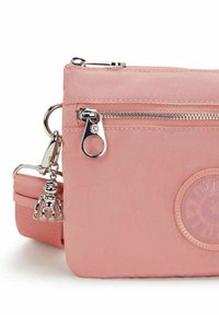 Růžová crossbody kabelka s kovovými prvky, zipovou kapsou a odnímatelným charms opice. Na přední straně se nachází kulaté logo.