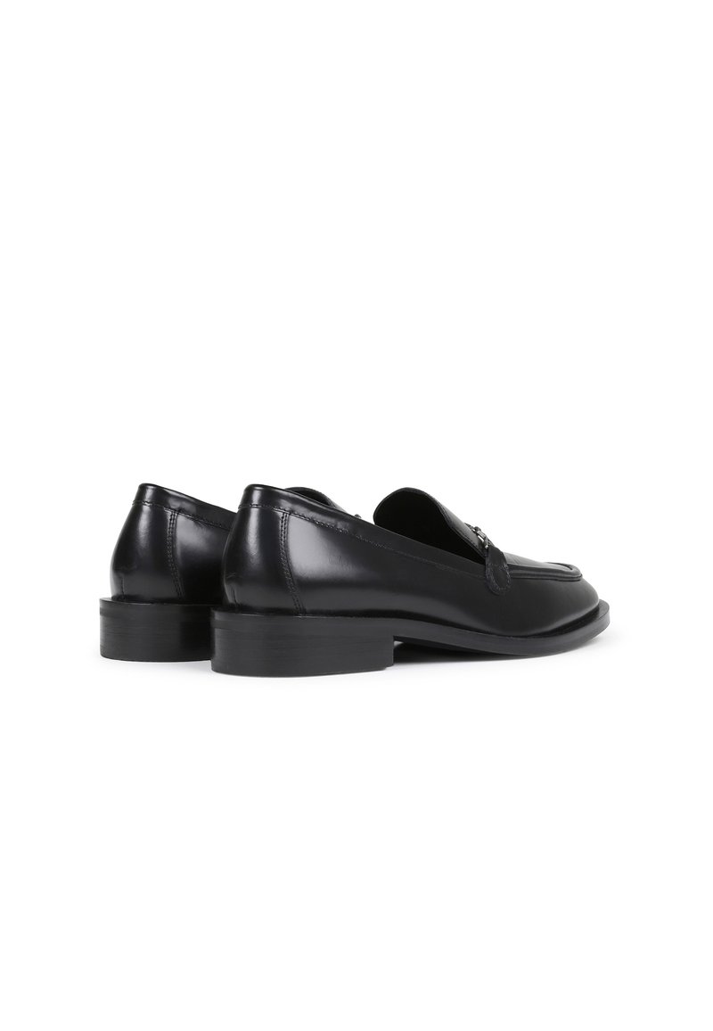 Mocassins en cuir noir avec une texture lisse, des talons plats et des coutures décoratives sur le dessus. Comprend un petit accent métallique pour le détail.