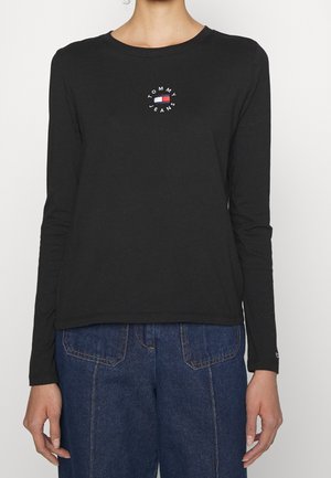 Nainen, joka pukeutunut mustaan pitkähihainen Tommy Jeans -paitaan, jossa rintakehässä keskitetty logo, yhdistetty korkeavyötäröisiin sinisiin farkkuihin.
