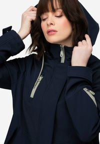 Chaqueta softshell de color azul marino con cuello alto, capucha ajustable y detalles en cremallera de color gris claro. Cuenta con dos bolsillos laterales y una tela texturizada.