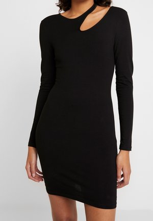 Femme portant une robe noire ajustée à manches longues avec un décolleté asymétrique et un détail découpé sur une épaule.