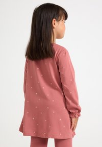Junges Mädchen mit glattem, dunklem Haar trägt ein langärmliges, staubrosa Kleid mit kleinen weißen Blumenmustern und steht seitlich vor einem weißen Hintergrund.