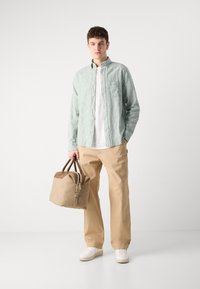 Camicia a righe verde e bianca a maniche lunghe sopra una maglietta bianca, abbinata a pantaloni beige. Porta una borsa marrone chiaro e marrone. Sneakers bianche.