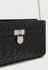 Mustast nahast käekott, millel on punutud tekstuur, hõbedased metalldetailid ja ümmargune logokaunistus. Omab kettidega rihma ja turvalist klambriga sulgurit.