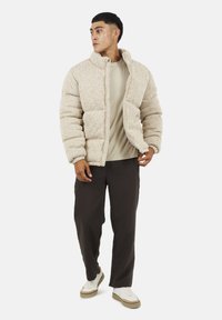 Giacca imbottita beige con tessuto testurizzato, colletto alto, chiusura a zip frontale, indossata sopra una camicia chiara e pantaloni scuri, abbinata a sneakers bianche.