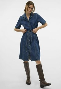 Denimklänning i mörkblått med korta ärmar, knapprad fram, bältesmidja och lätt utsvängd kjol, kombinerad med höga bruna stövlar.