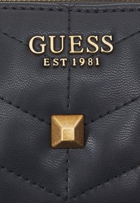 Černá syntetická peněženka s prošívaným designem, zlatými kovovými doplňky a čtvercovou kovovou ozdobou. Text uvádí "GUESS EST 1981."