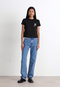 Camiseta de algodón negra de manga corta con un pequeño gráfico blanco en el pecho, combinada con jeans azules de pierna recta y zapatos negros.