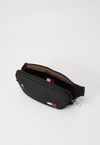 Zwarte stoffen heuptas met ritsluiting, bruin interieur en een klein logopatch met rode, witte en blauwe accenten.