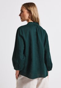 Dark green, long-sleeve blouse with a relaxed fit. Features a ruffled neckline and buttoned cuffs. Smooth texture and slightly curved hem.