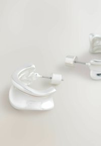 Sudraba metāliskas auskari ar viļņotu, abstraktu formu un spīdīgu virsmu. Aprīkoti ar āķveida aizdari drošai nēsāšanai.