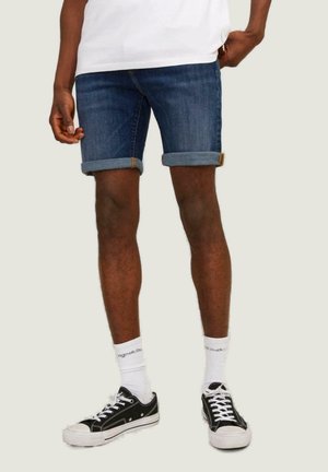 Shorts de mezclilla con un lavado azul oscuro, puños doblados, bolsillos, combinados con zapatillas negras y calcetines blancos hasta el tobillo. Diseño simple y ajuste casual.