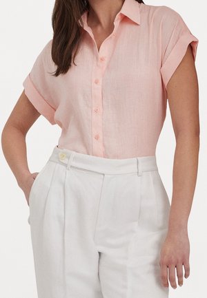 Femme portant une chemise rose clair à manches courtes boutonnée, rentrée dans un pantalon blanc taille haute avec poches latérales.