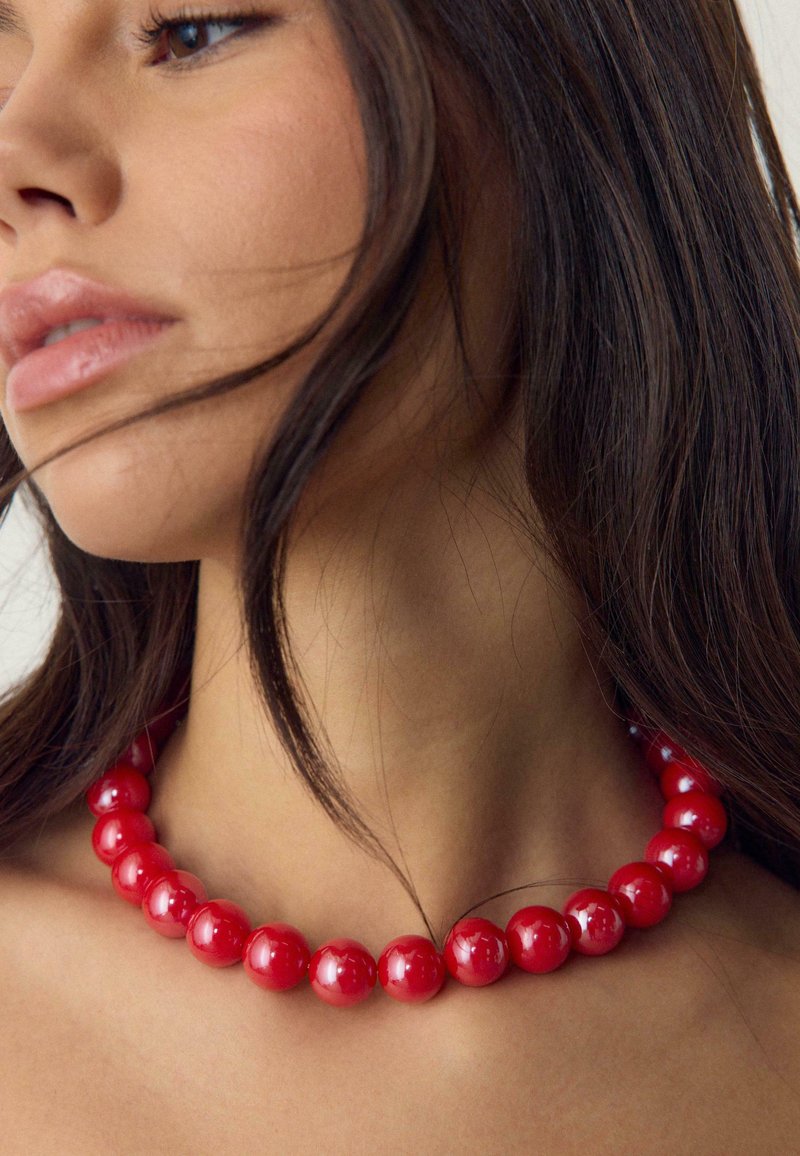 Gros plan sur une femme portant un collier de perles rouges brillantes, avec des cheveux foncés partiellement couvrant son visage et son cou.