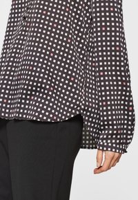 Svart och rosa rutig långärmad blus med manschettärmar, bärs över svarta byxor, och visar den nedre överkroppen och högerhanden.